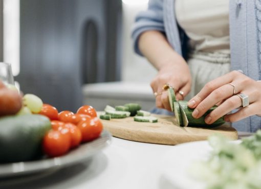 Close-up of a woman slicing cucumbers on a wooden board surrounded by fresh vegetables.